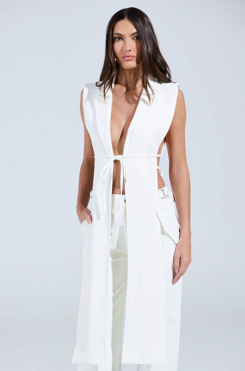 A person with long hair wears the AKIRA Label HONEY HONEY TIE VEST, characterized by a high-low silhouette and waist tie, paired with matching pants. The look is elegant against a plain light-colored background.