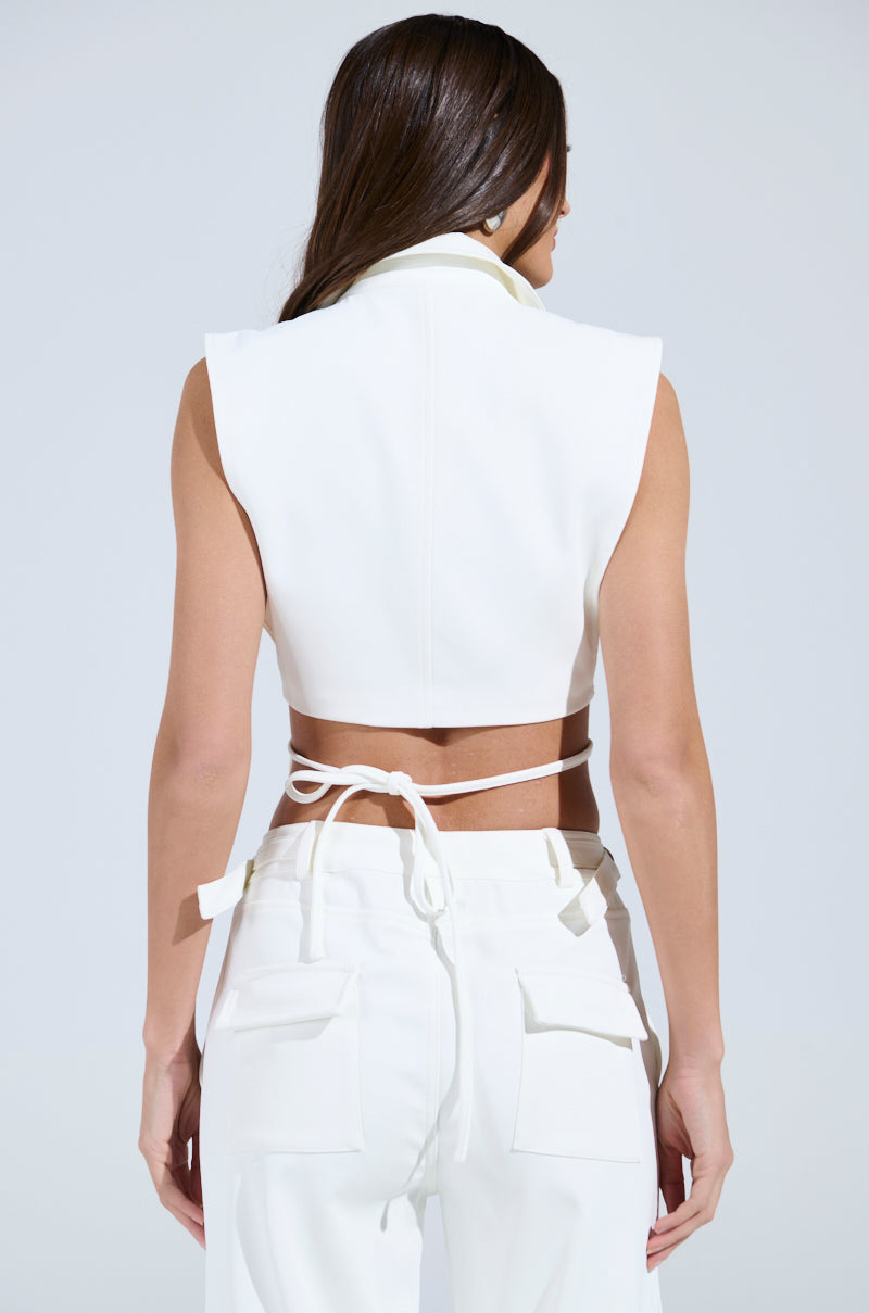 A person with long hair is seen from behind in a HONEY HONEY TIE VEST sleeveless white crop top, featuring a tie detail at the back, with white high-waisted pants against a light gray background.