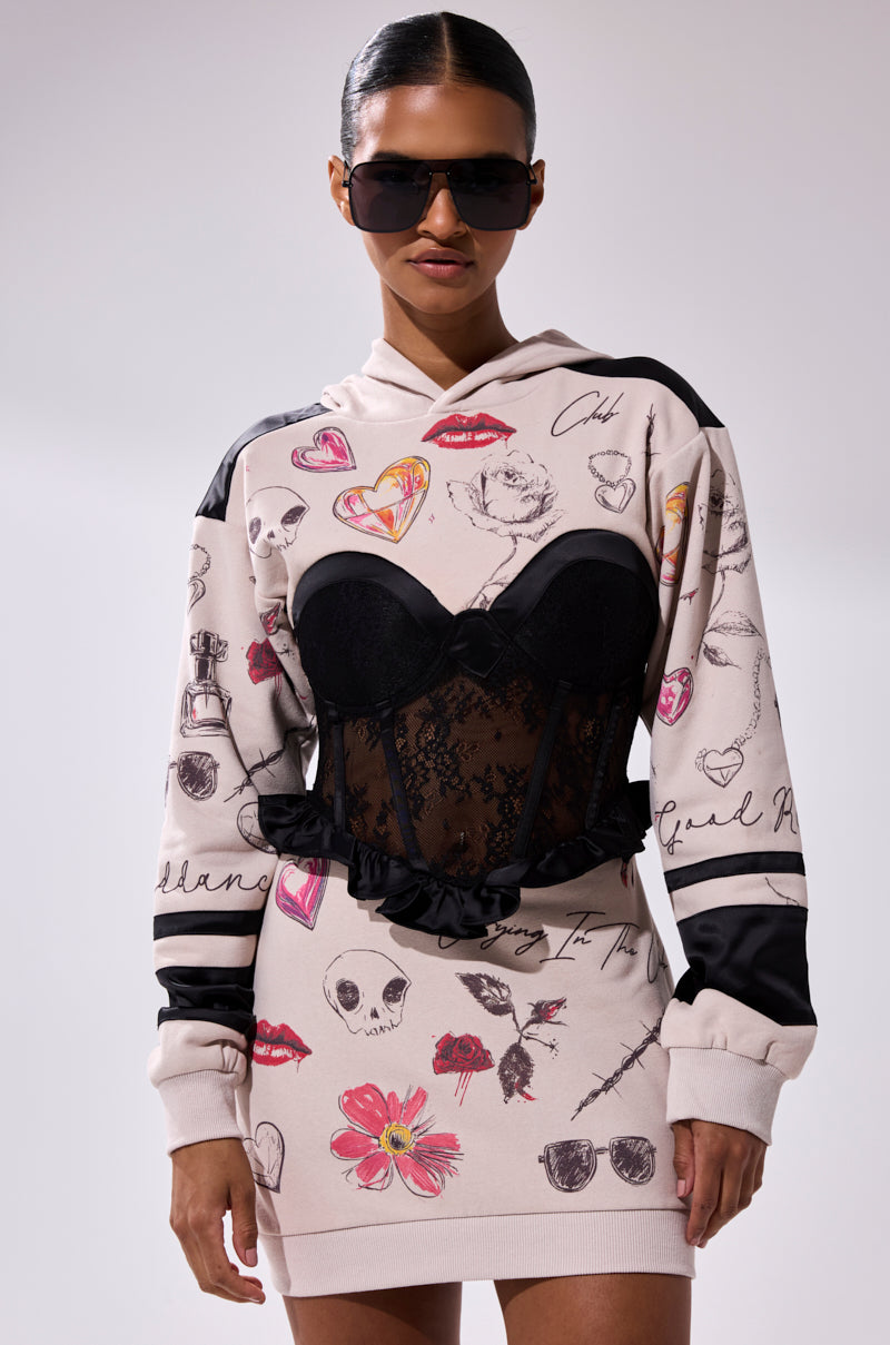 A woman in the SNATCH ME UP CORSET SWEATSHIRT DRESS—featuring large black sunglasses, an oversized fit with heart, skull, and lipstick print sketches, and a black lace corset overlay—stands against a plain background.