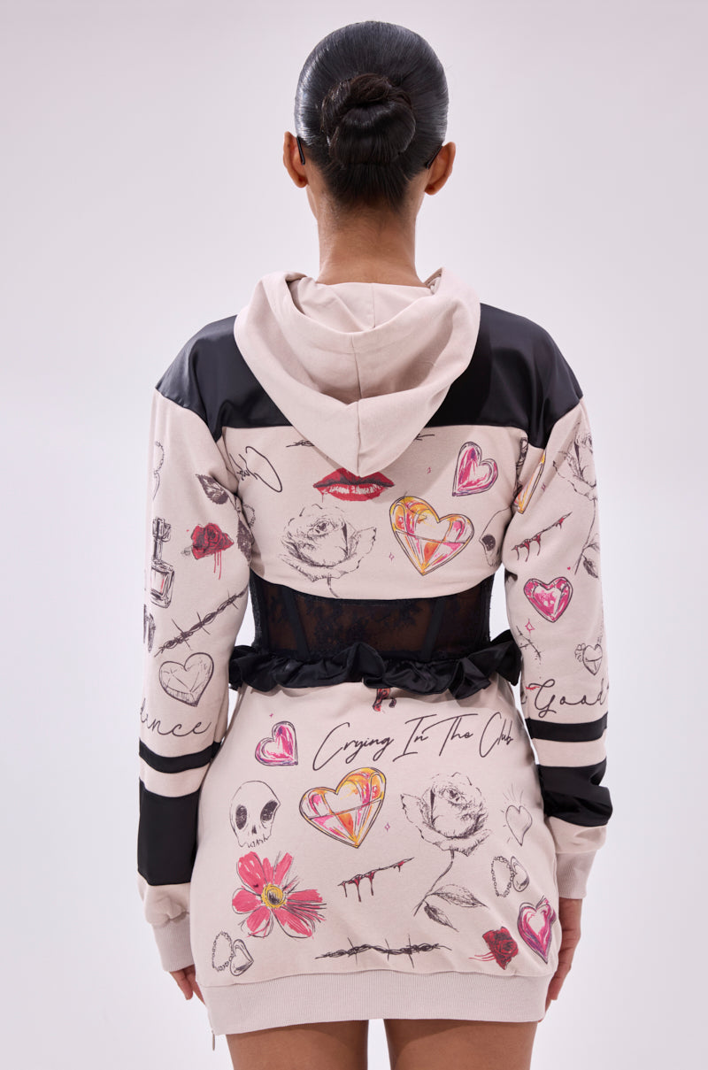 A woman with a sleek bun faces away, wearing the SNATCH ME UP CORSET SWEATSHIRT DRESS—an oversized hoodie dress with black lace at the waist and a colorful hand-drawn print of hearts, lips, flowers, skulls, and text on a light background.