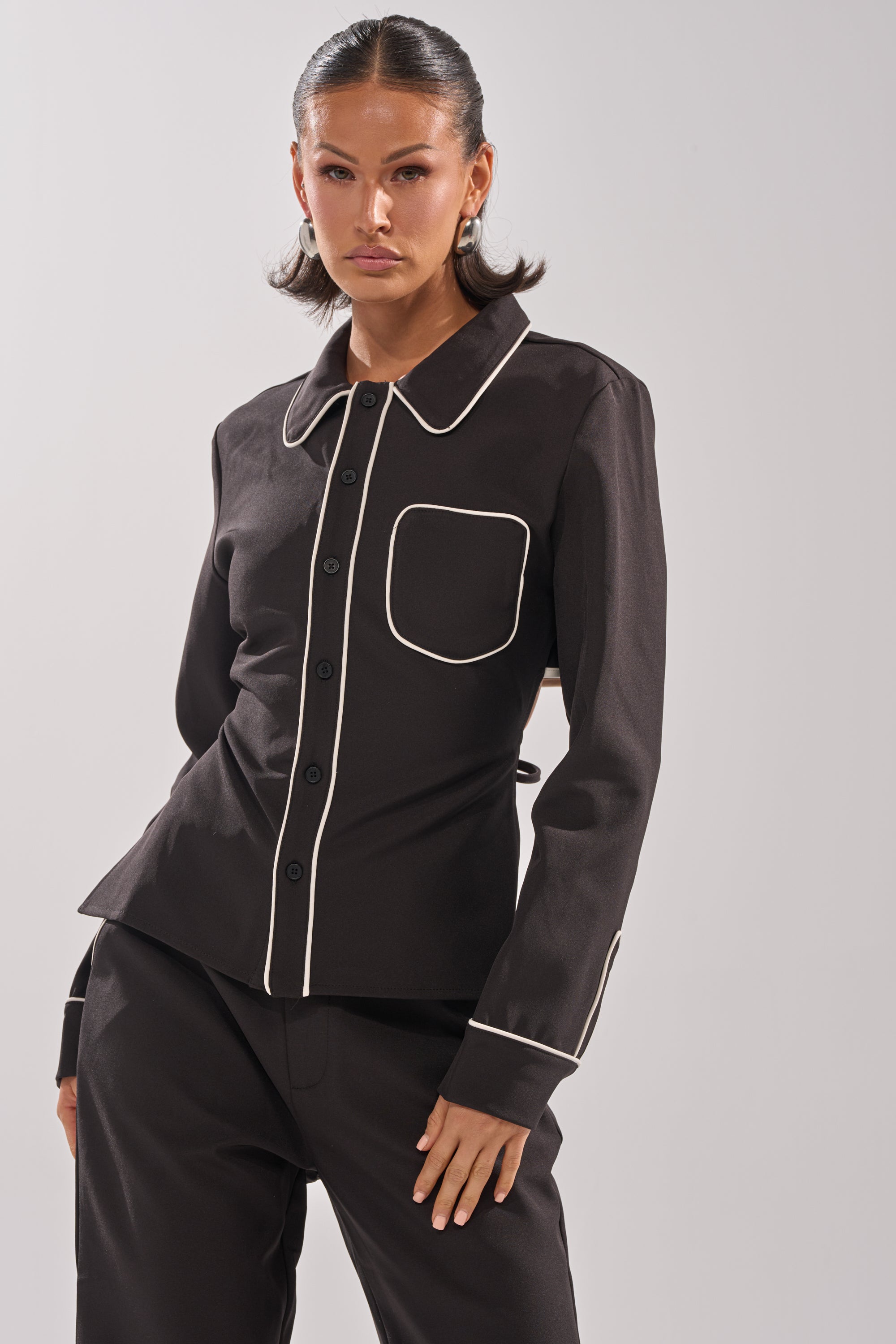 Someone stands against a plain light background wearing the PARTY IN THE BACK BUTTON DOWN blouse, featuring white piping on the collar, pocket, and cuffs, paired with matching black pants.