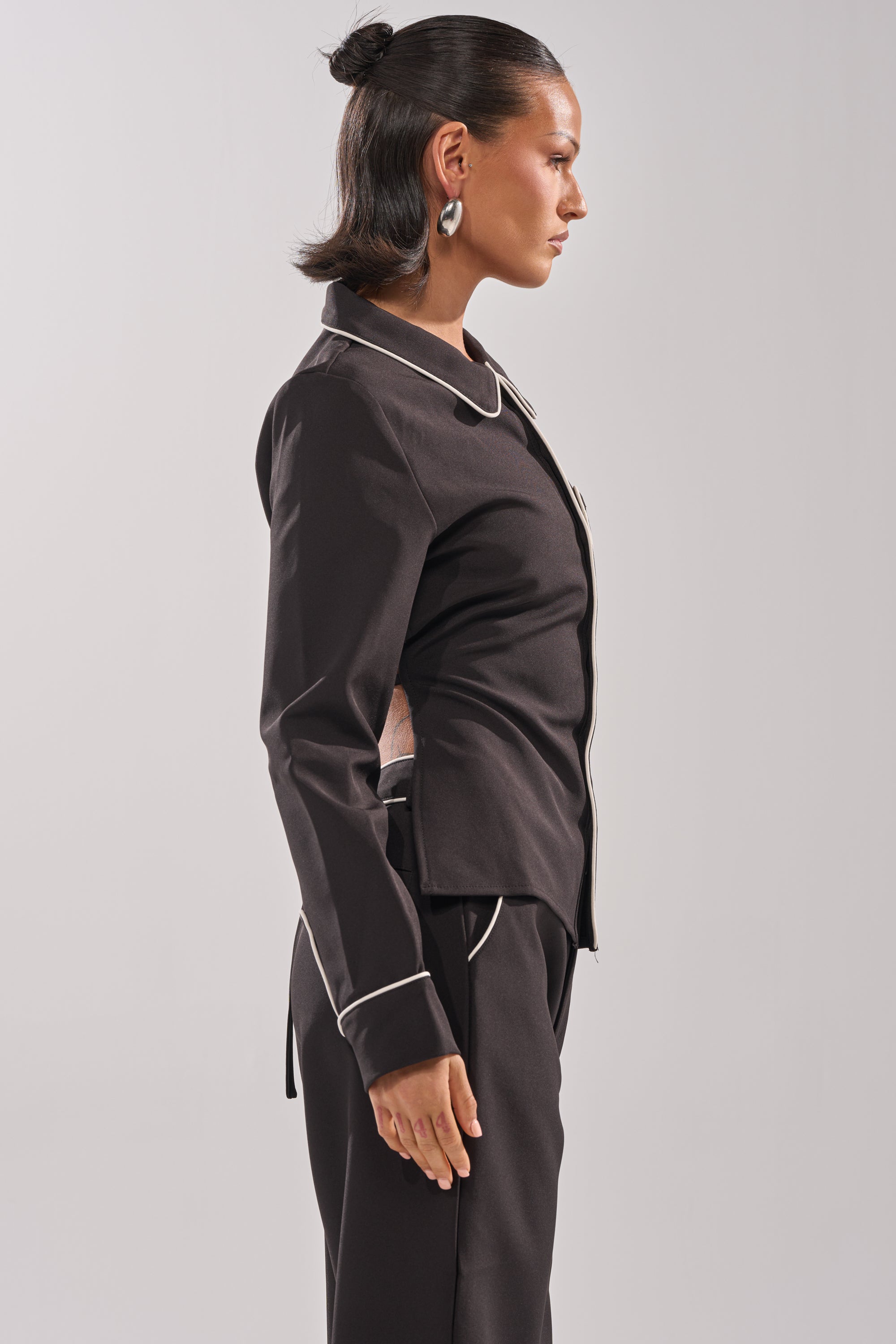 A woman in profile models the PARTY IN THE BACK BUTTON DOWN, a fitted black shirt with white piping on the collar and cuffs, paired with matching pants. She stands against a light gray background, her hair in a sleek bun and wearing large round earrings.