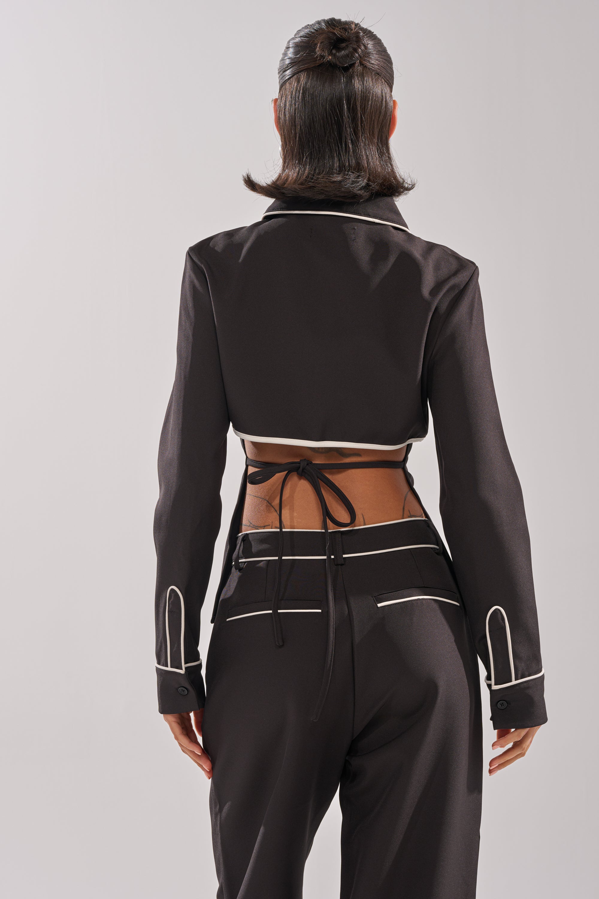 A woman with short, dark hair is seen from behind wearing the PARTY IN THE BACK BUTTON DOWN, a black tie-back shirt with white trim and matching high-waisted pants. The shirt exposes her lower back and ties with thin strings against a light background.