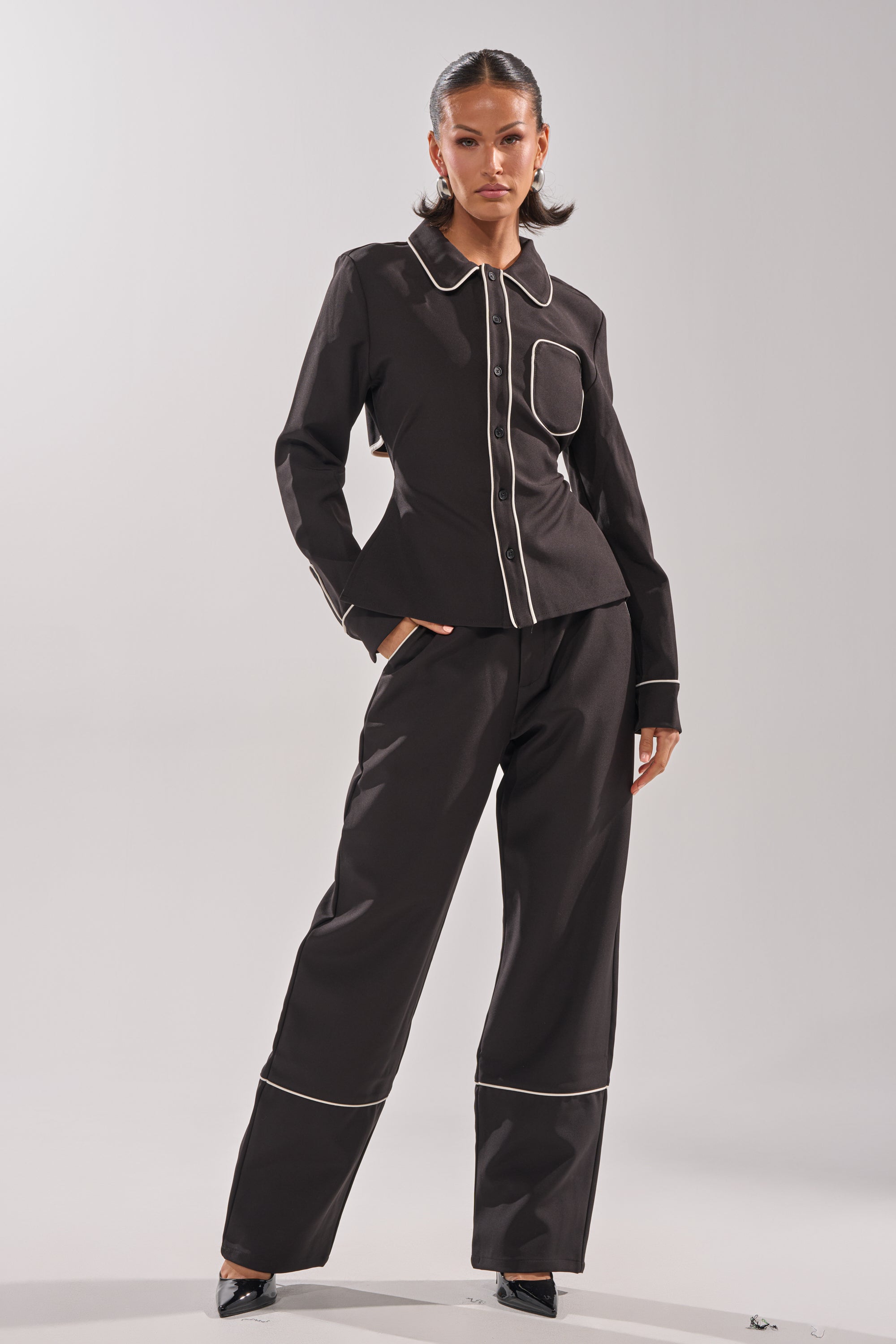 A woman exudes confidence in the PARTY IN THE BACK BUTTON DOWN blouse, paired with a black pantsuit featuring white piping on the collar, cuffs, and hems. With slicked-back hair and black pointed heels, she stands against a plain background.