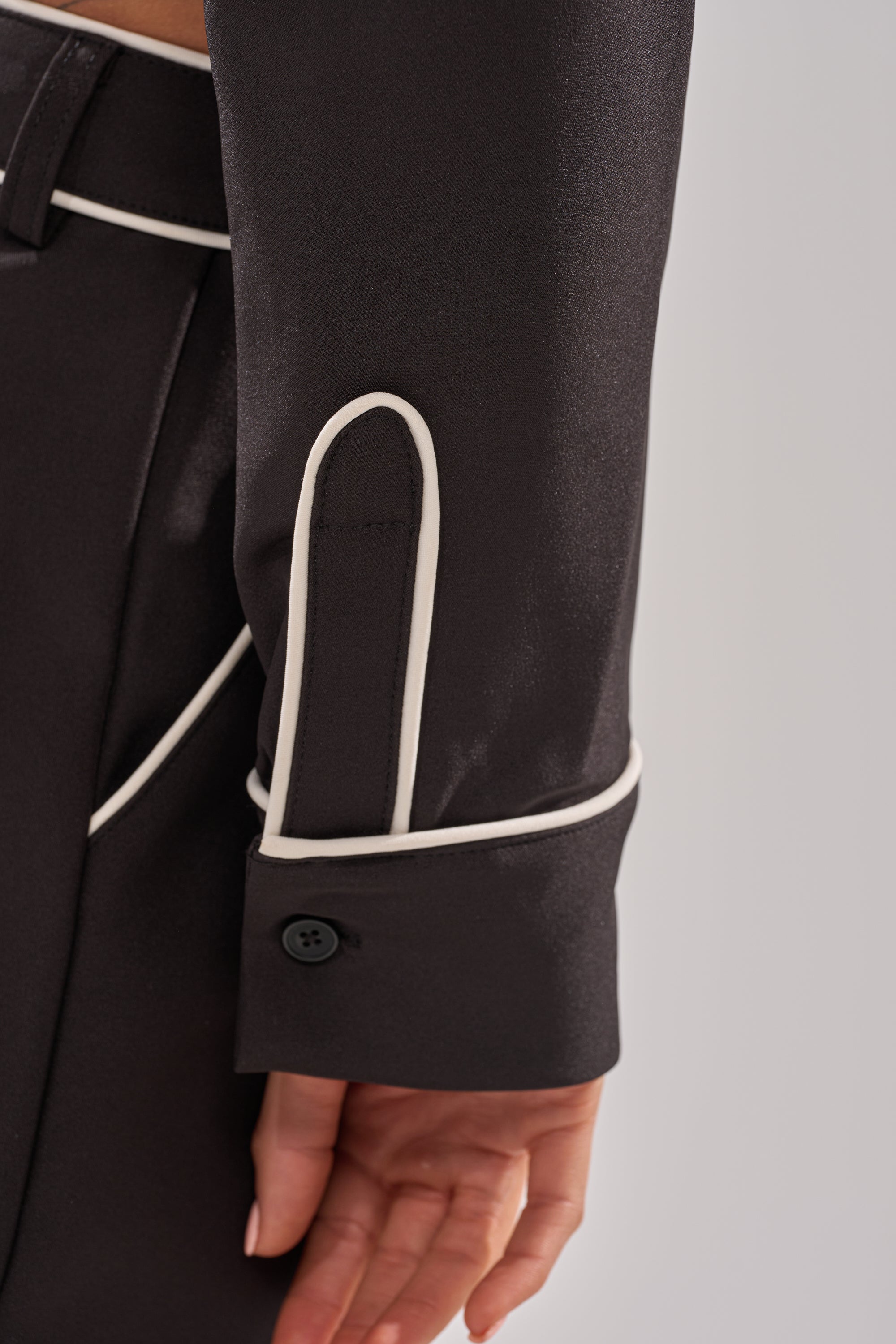 A close-up of a person’s arm wearing the PARTY IN THE BACK BUTTON DOWN—black satin blouse with a tailored silhouette, white piping on the sleeve and pocket, and a black button at the cuff.