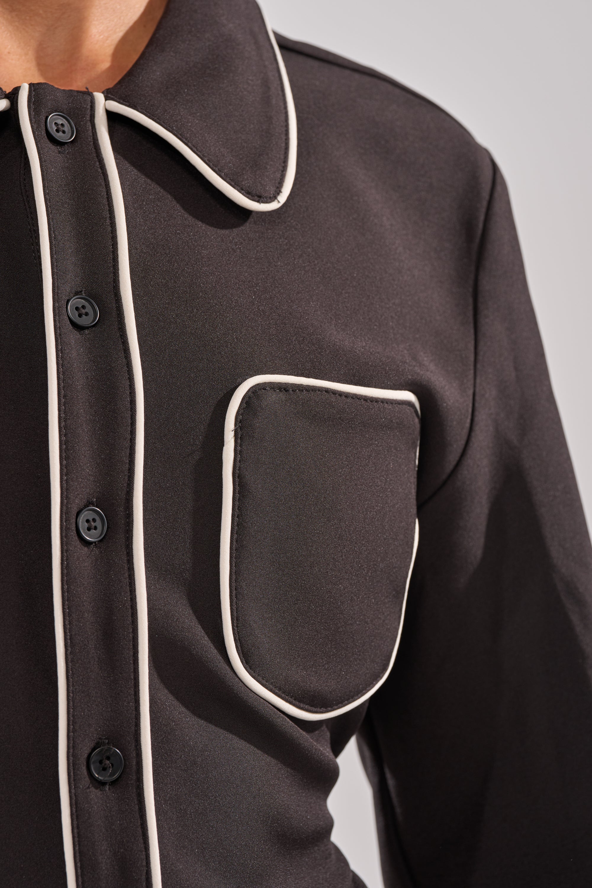 A close-up of the PARTY IN THE BACK BUTTON DOWN, a black tailored blouse featuring white piping on the collar, front placket, and chest pocket, worn by a person against a plain light background.