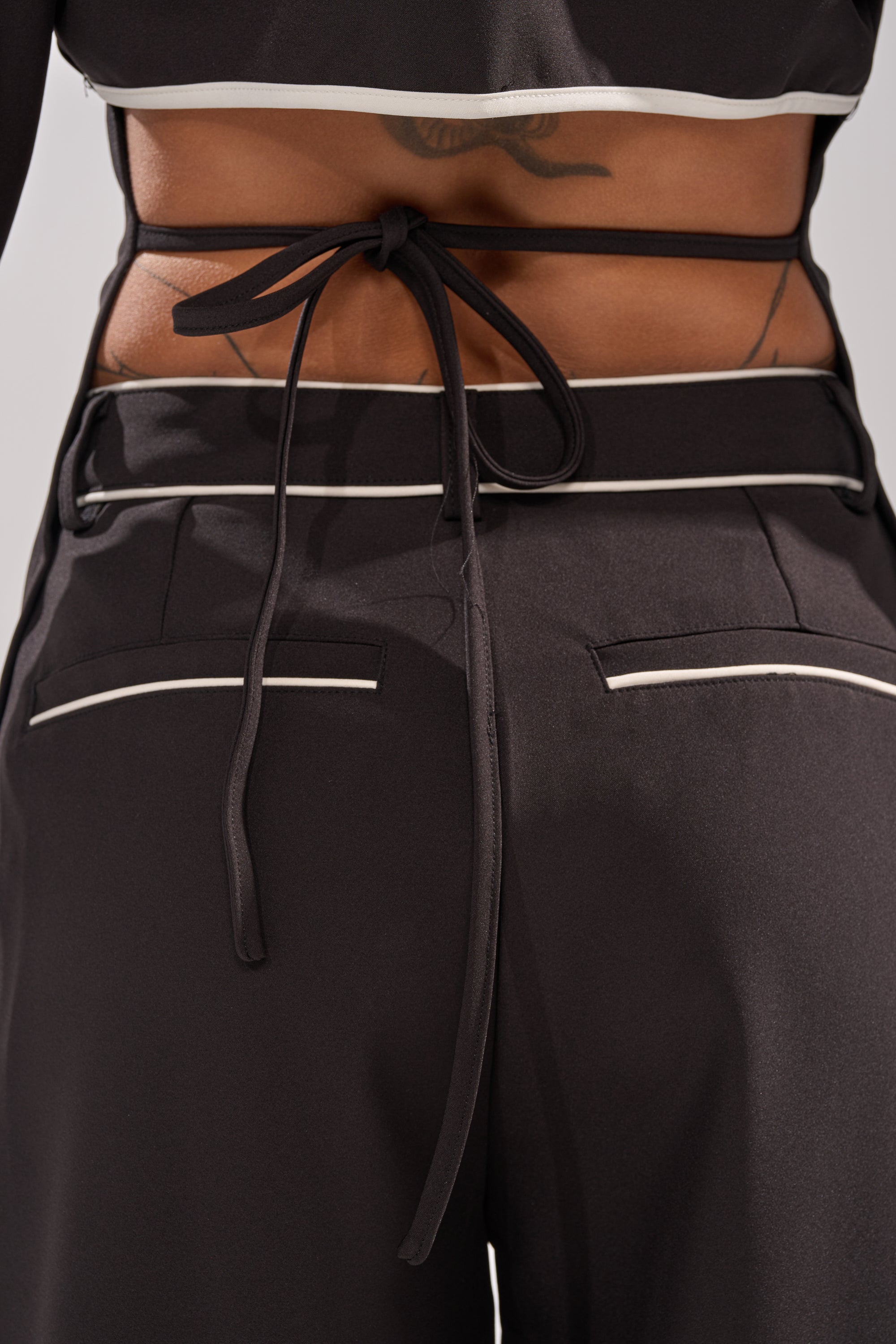 Close-up of a person’s lower back wearing the PARTY IN THE BACK BUTTON DOWN, a black tailored blouse with white piping and a bow at the back, partially revealing a tattoo above black pants’ waistband.