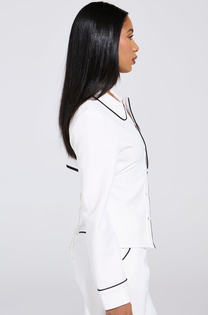 A person with long, straight black hair is standing in profile against a plain white background, wearing a white suit with black trim over the LISA WOVEN PIPED BUTTON UP.