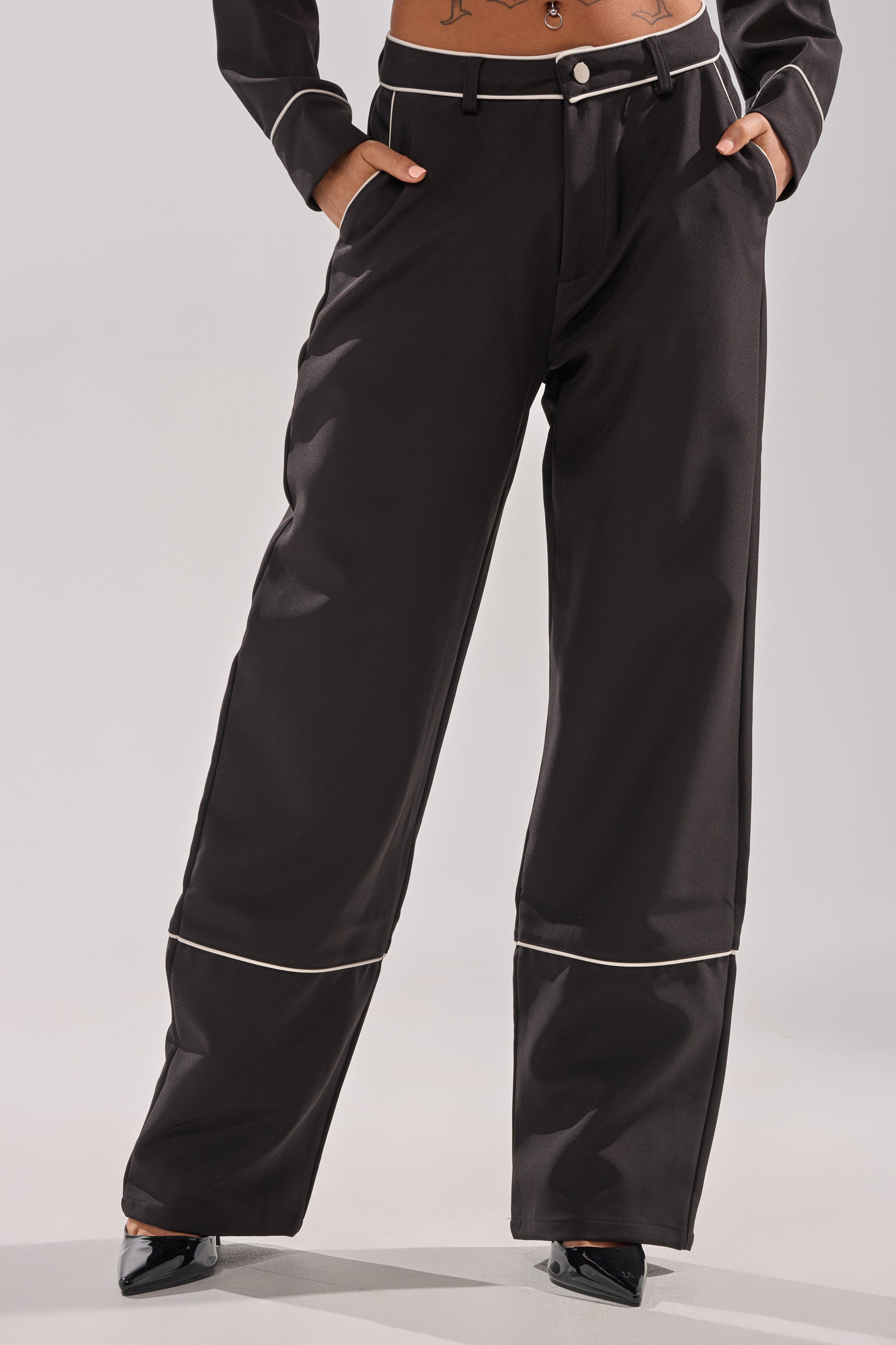 A person wearing PARTY AT MY PLACE TROUSER, featuring high-rise wide-leg fit with white piping, paired with black pointed-toe heels; hands in pockets, torso partially visible.