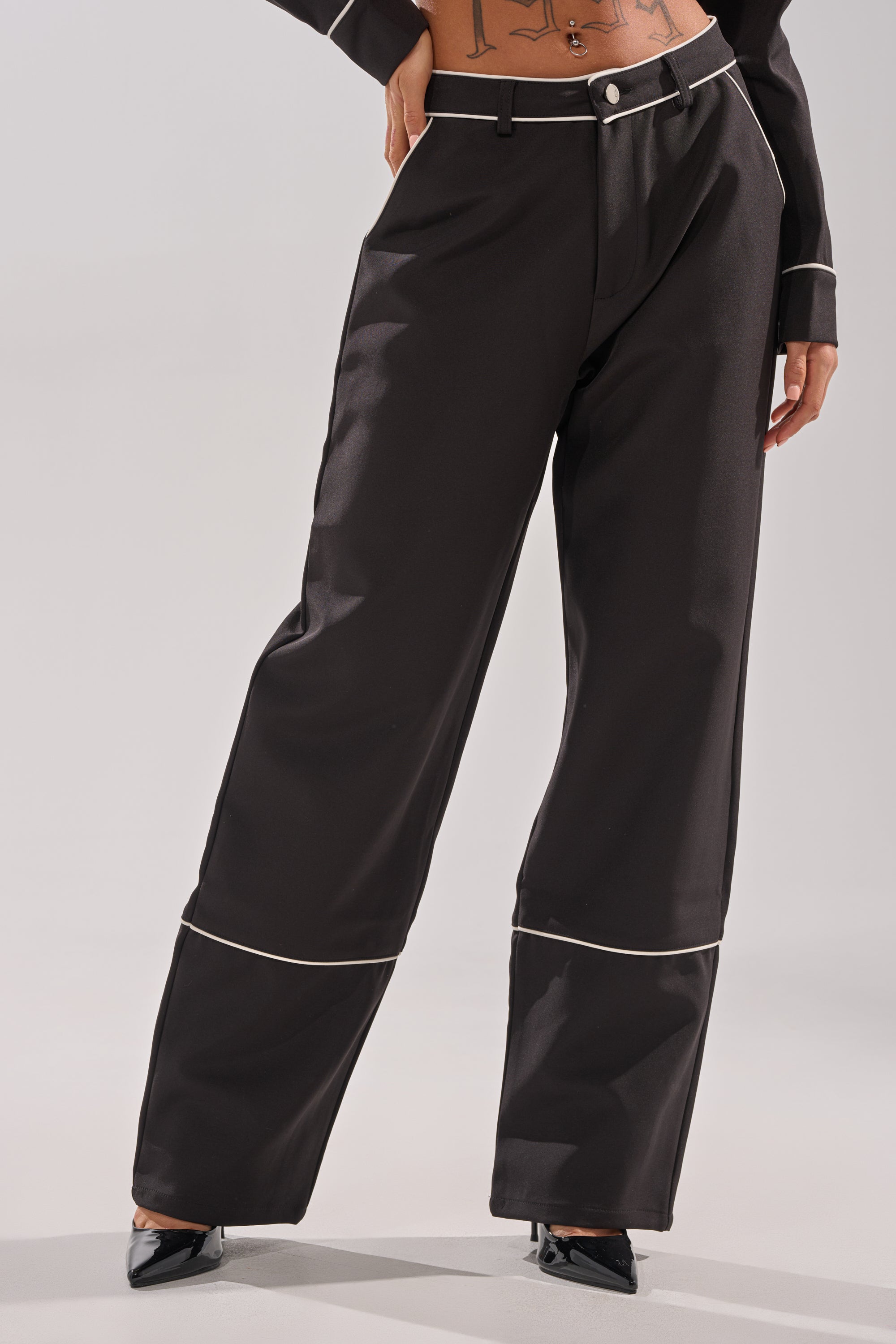 A person wears the AKIRA Label PARTY AT MY PLACE TROUSER, high-rise wide-leg style with white piping along seams and cuffs, paired with black pointed-toe heels. Their midriff is exposed, showing a tattooed torso.
