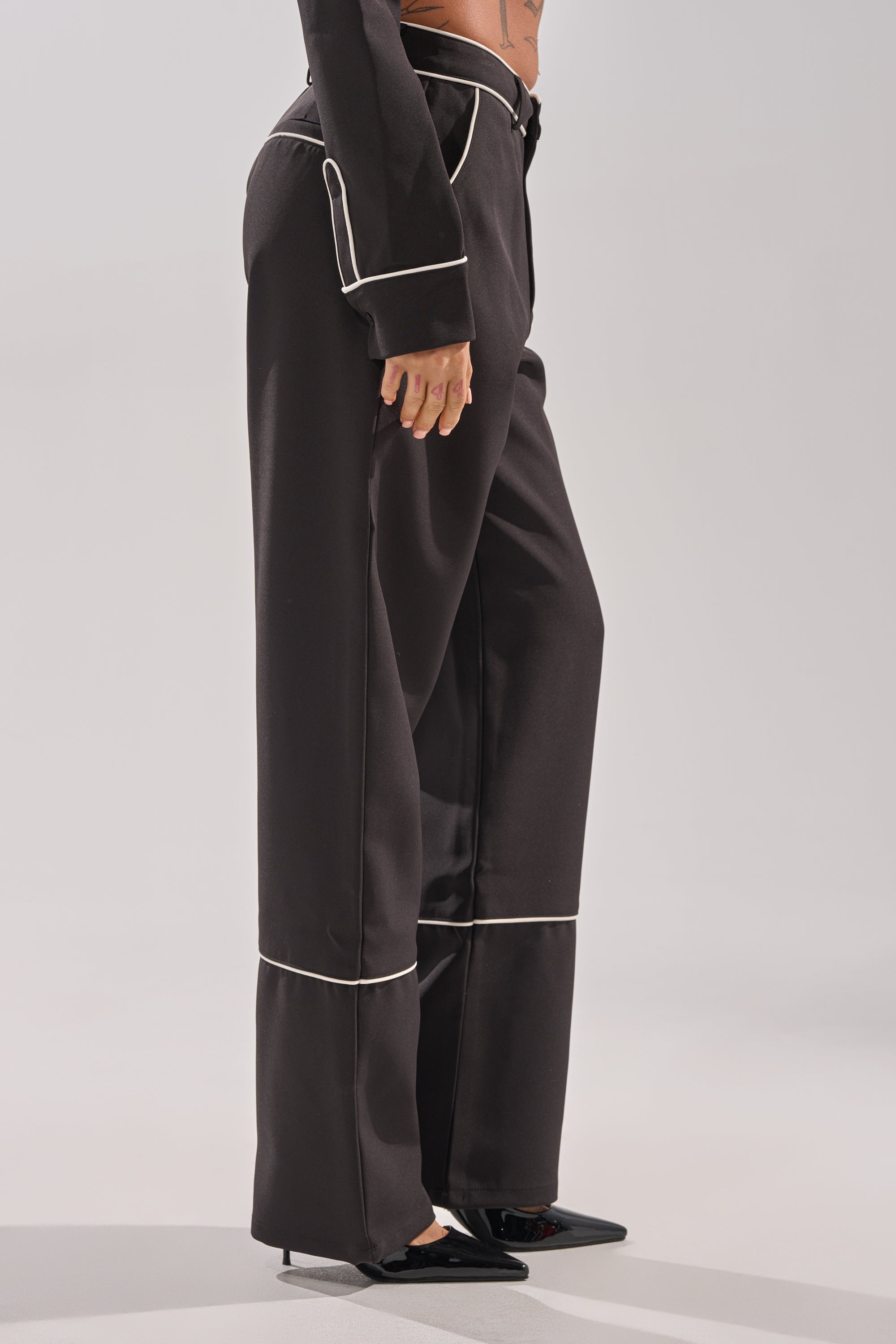A person wears the PARTY AT MY PLACE TROUSER—high-rise, wide-leg pants with white piping—and black heels, standing on a light background. Only the lower half is shown, drawing attention to these stylish party trousers.