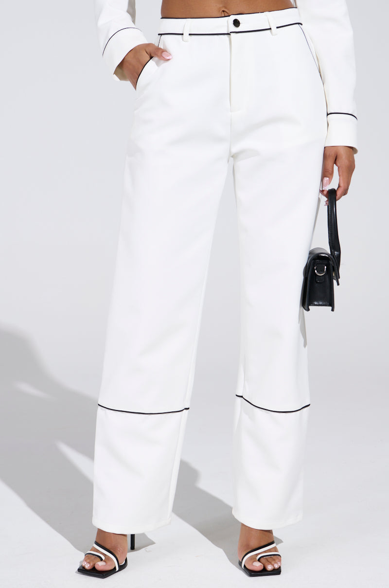 A person in LISA CUFFED WOVEN PANT with black contrast piped detailing, paired with a matching cropped jacket and black heeled sandals. They hold a small black handbag against a neutral background.