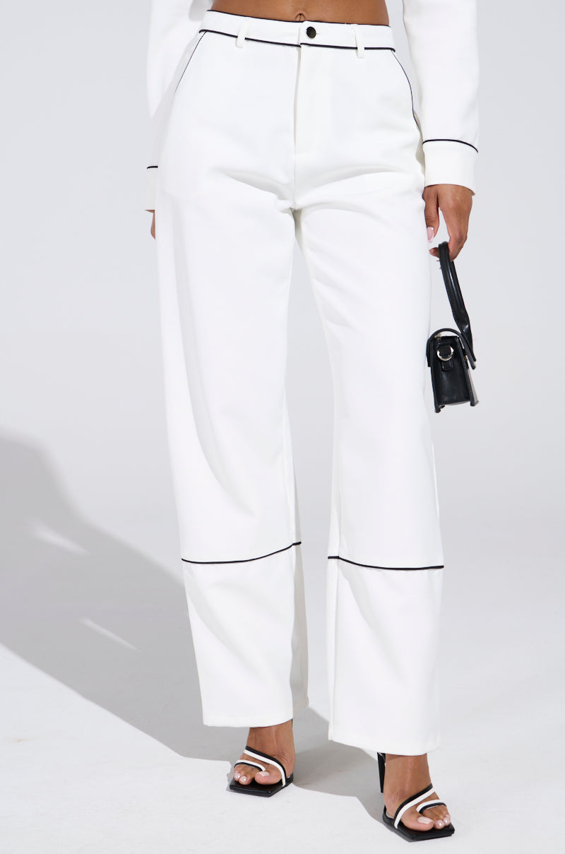 A person wears LISA CUFFED WOVEN PANT in white with black piping, paired with a matching top. They hold a small black purse and wear black and white open-toe heels, creating a striking look against the plain light gray background.