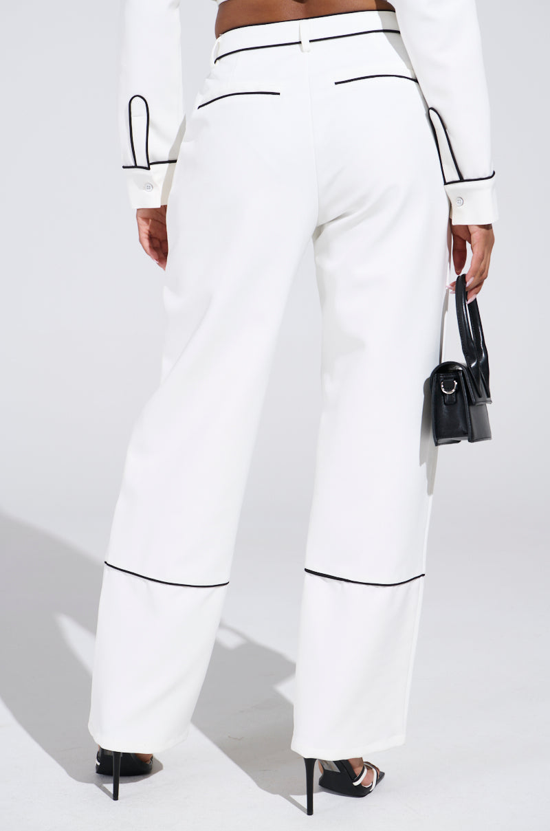 A person wears LISA CUFFED WOVEN PANT in white and a matching jacket, both with contrast piped detailing. They're accessorized with a small black handbag and black high heels against a plain white background.