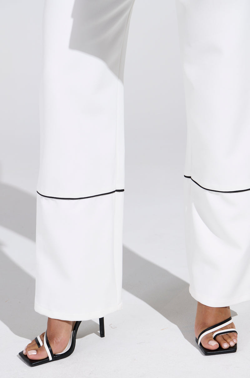 A person's lower legs are visible in LISA CUFFED WOVEN PANT, featuring black contrast piped detailing, paired with black high-heeled sandals. The background is a plain white surface with shadows cast by the legs.