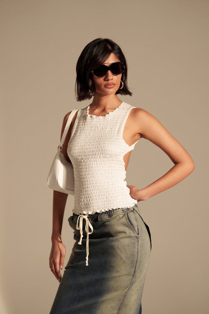 A woman wearing sunglasses and the CHLOE OPEN BACK & SMOCKED BODY TOP, styled with a long denim skirt and small white shoulder bag, poses confidently with one hand on her hip against a plain beige background.