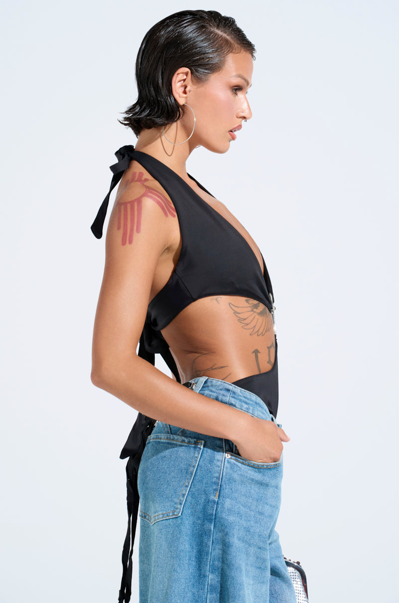A person with short hair wears the sleeveless black LOTS TO GIVE BODYSUIT with a plunging neckline and light blue jeans, showcasing arm and side tattoos. Facing sideways, they sport hoop earrings against a plain light gray background. 4