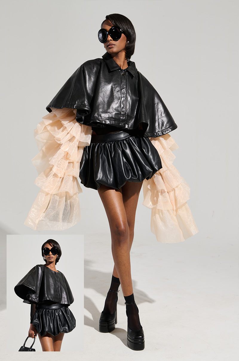 A model exudes confidence in the LOUISE TULLE SLEEVE CAPE, a black faux leather ensemble featuring dramatic puffy tulle sleeves and a chic cropped silhouette. Her oversized sunglasses and platform shoes complement the look, while she holds a small handbag. The simple background accentuates her avant-garde style. 0