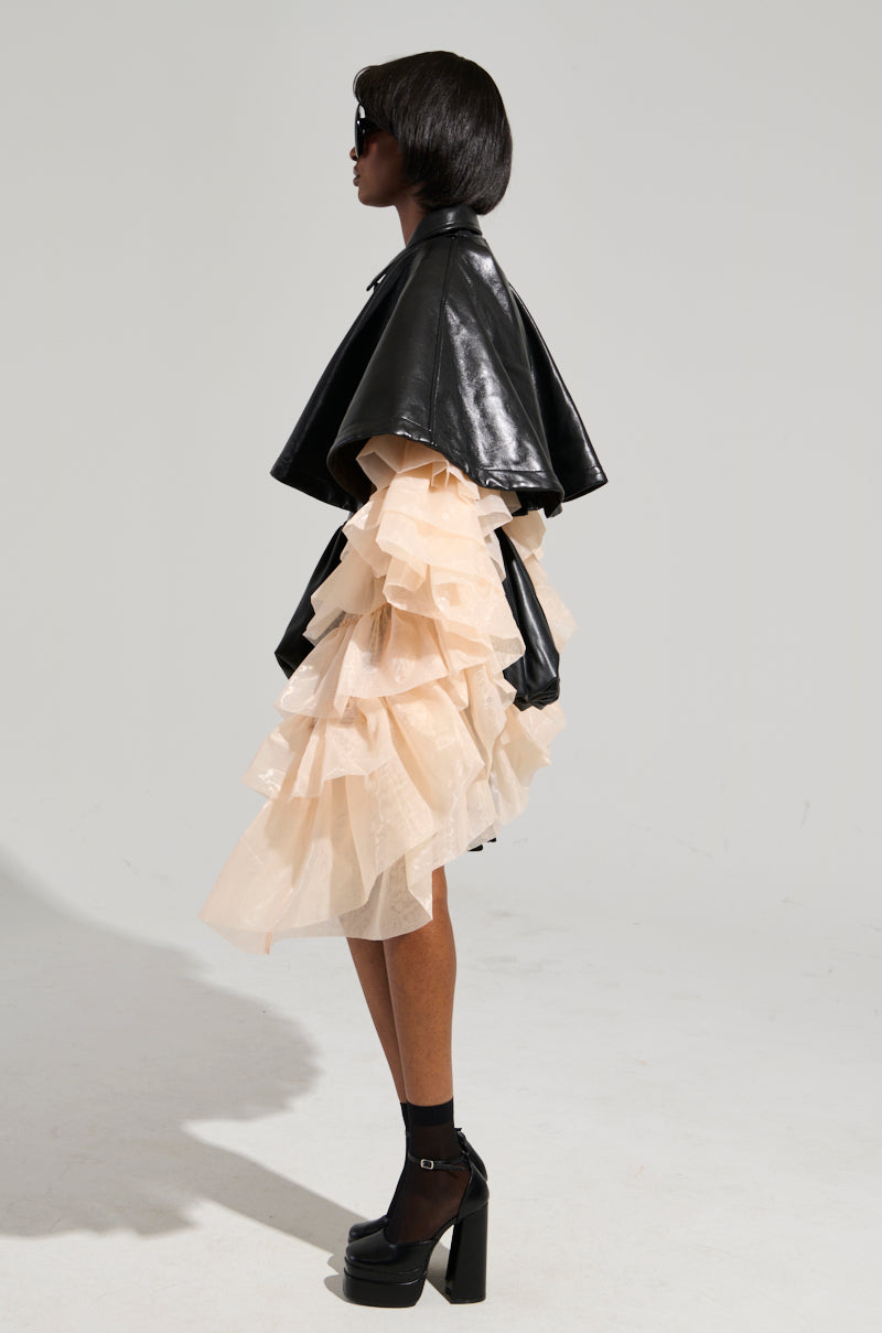 A person stands in profile wearing the stylish LOUISE TULLE SLEEVE CAPE over a cream-colored, ruffled dress. They have short hair, sunglasses, and are sporting black platform heels with socks against a plain, light-colored background. 9