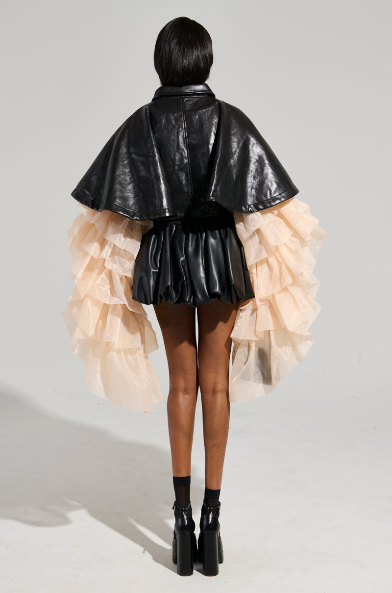 A person stands with their back turned, showcasing a cropped silhouette adorned with a black leather jacket and short skirt. The LOUISE TULLE SLEEVE CAPE, made of beige, ruffled fabric, drapes elegantly from the sides like a faux leather cape. They complement the look with black high-heeled shoes and have short, dark hair, projecting an effortlessly chic vibe. 10