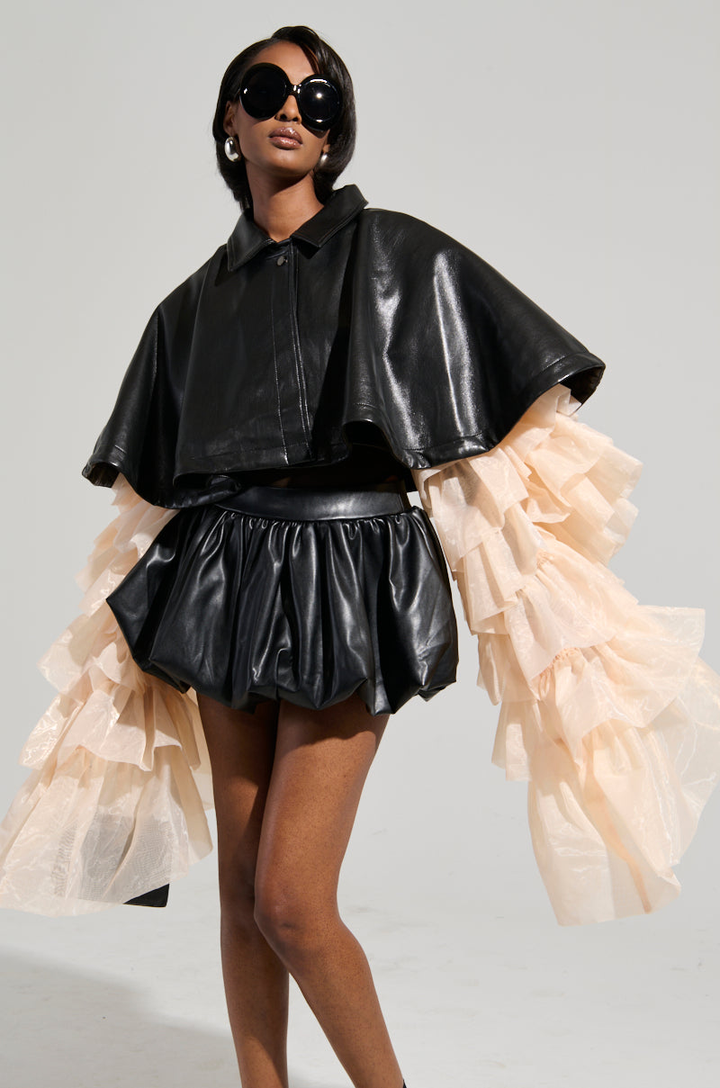 An elegant person dressed in the LOUISE TULLE SLEEVE CAPE, featuring oversized black sunglasses, a cropped silhouette with a faux leather cape, and a matching high-waisted skirt adorned with puffy, layered tulle sleeves stands against a plain background. The ensemble exudes boldness and chic style. 1