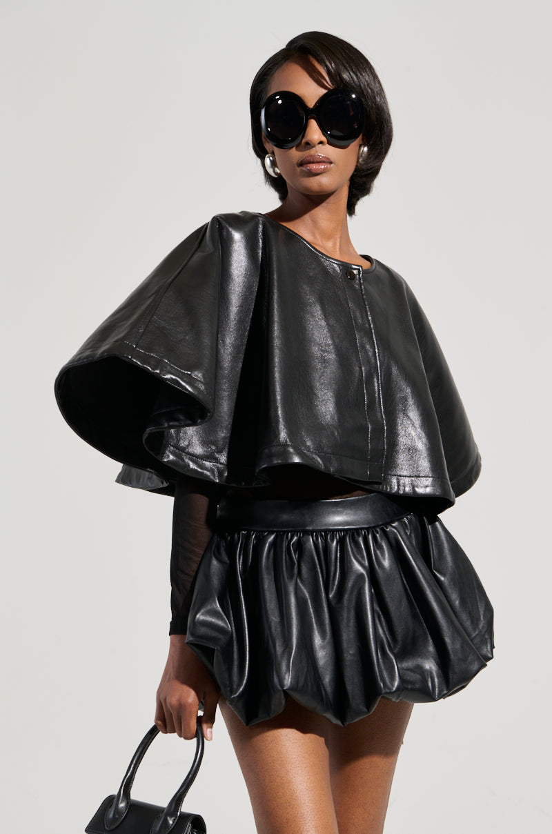 A woman wearing oversized black sunglasses and a chic leather outfit, featuring the LOUISE TULLE SLEEVE CAPE with a puffy skirt, exudes confidence. She holds a black handbag, her ensemble elevated by the cropped silhouette of her top against the plain backdrop. 3