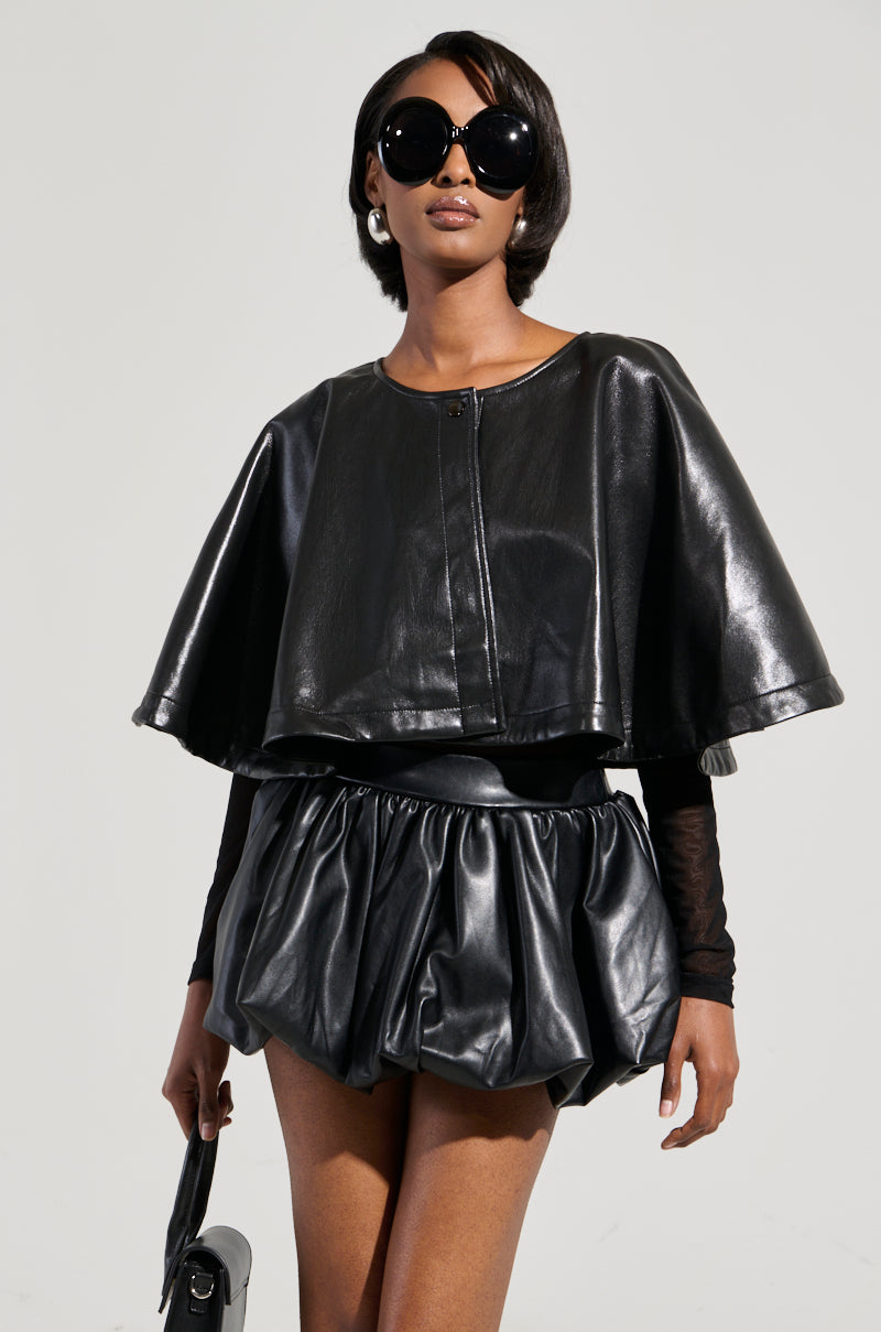 A person wearing oversized sunglasses and a black leather ensemble, featuring the LOUISE TULLE SLEEVE CAPE and a puffed mini skirt. They're standing against a plain background, holding a small black handbag. 4