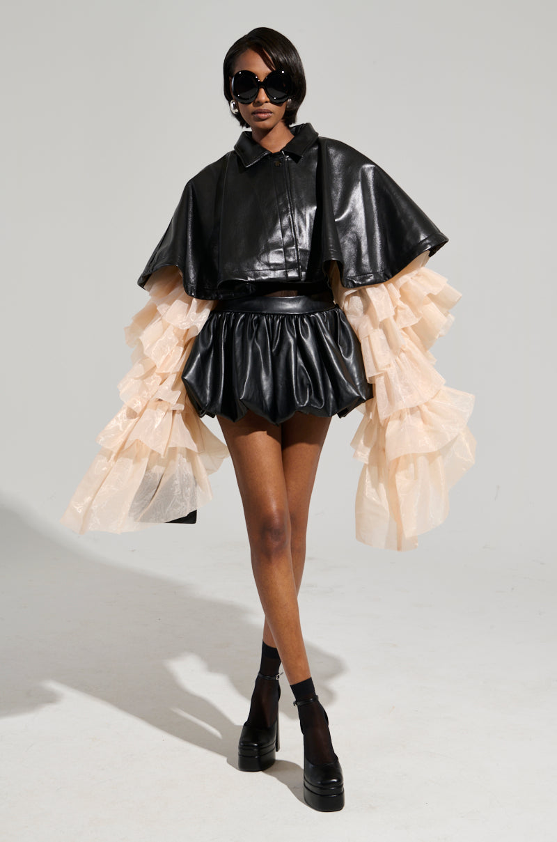 A model confidently poses in the LOUISE TULLE SLEEVE CAPE, featuring puffy sleeves and a tiered beige tulle underlay. She accessorizes with large sunglasses and platform shoes, showcasing her chic style against a plain background. 7