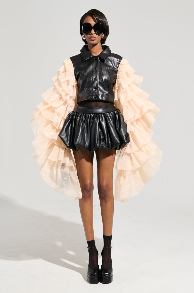 Wearing the LOUISE TULLE SLEEVE CAPE in a sleek cropped leather design paired with a mini skirt, this person exudes confidence. The dramatic beige tulle sleeves add flair to their ensemble, which is complemented by large sunglasses and chunky black platform shoes against a minimalist backdrop, creating an effortlessly chic look. 8