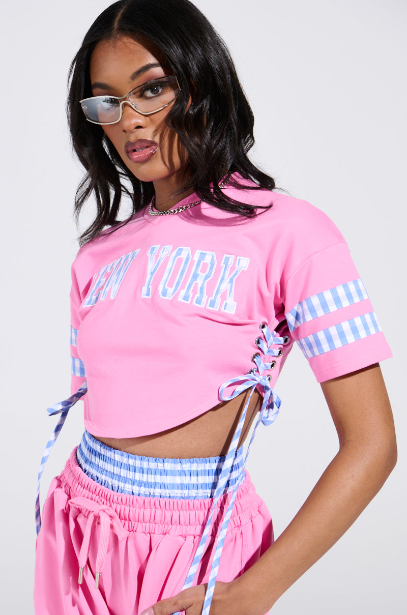 A person poses wearing a chic AKIRA Label NEW YORK GINGHAM CROP TOP featuring side lacing and a v-neckline. They pair it with matching pants and stylish sunglasses, while their long, loose hair enhances the sleek look.