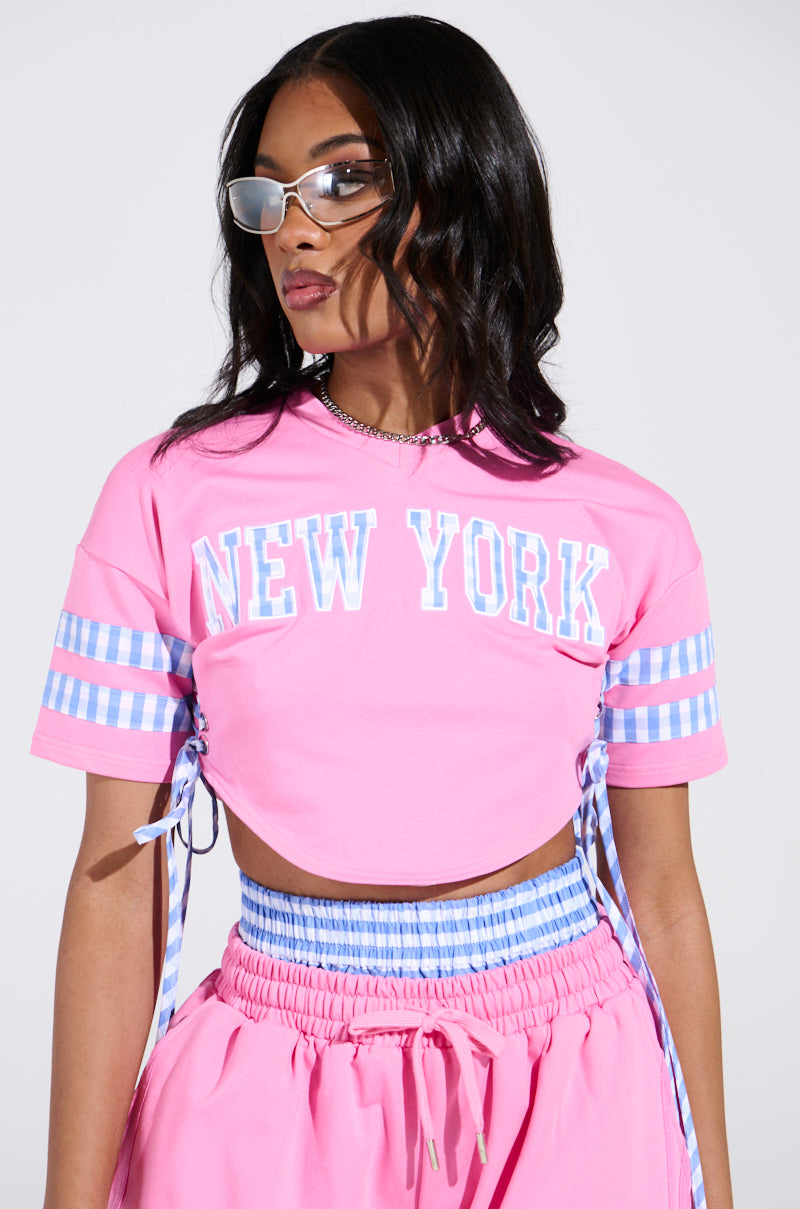 A woman dons a pink ensemble featuring an AKIRA Label NEW YORK GINGHAM CROP TOP with blue and white checkered stripe sleeves. She has long dark hair, sunglasses, and poses against a light gray background.