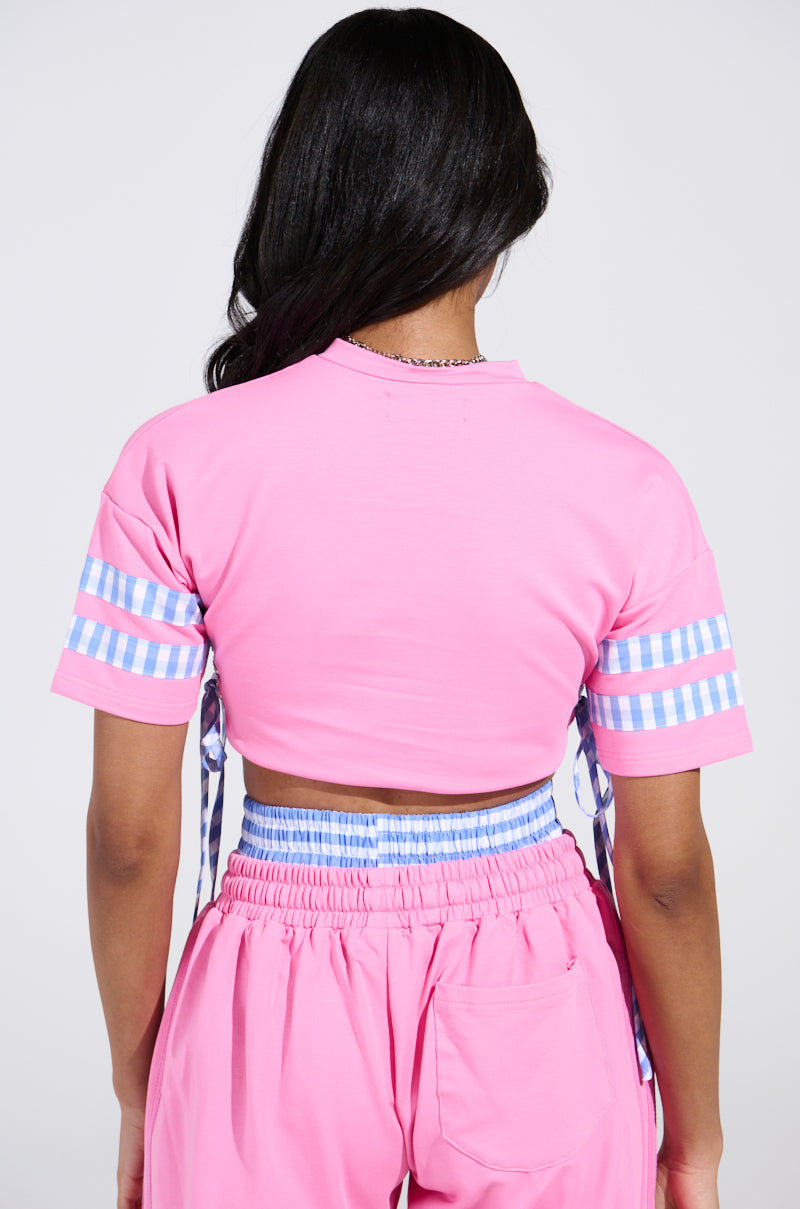 A person with long hair is wearing a pink NEW YORK GINGHAM CROP TOP featuring checkered blue and white sleeves, paired with matching shorts, while facing away from the camera.