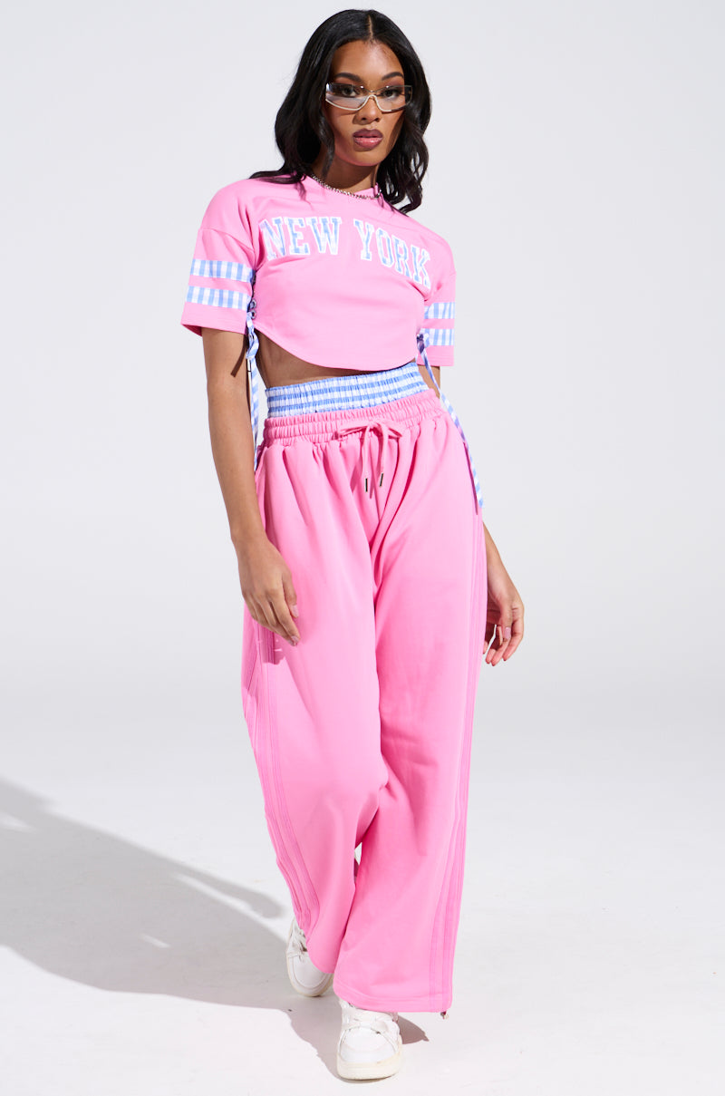 The woman confidently walks on a neutral background, wearing an AKIRA Label pink NEW YORK GINGHAM CROP TOP. The top has blue and white striped accents and a V-neckline, paired with pants that feature a drawstring waist. Her long hair cascades gracefully as she sports glasses.