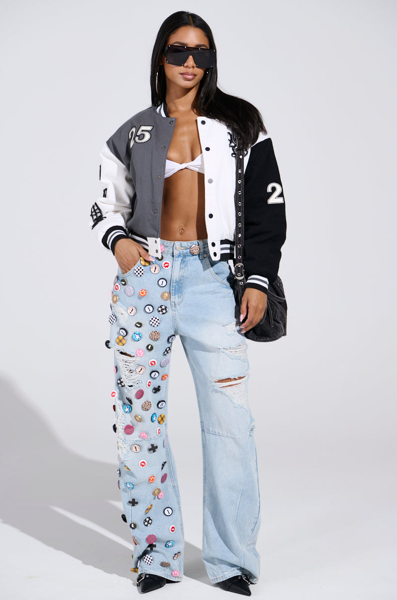 A woman in oversized sunglasses wears the NOT YOUR BABE EMBROIDERED BOMBER over a white bikini top, paired with light blue jeans featuring colorful patches. She holds a black handbag and stands against a plain light background.