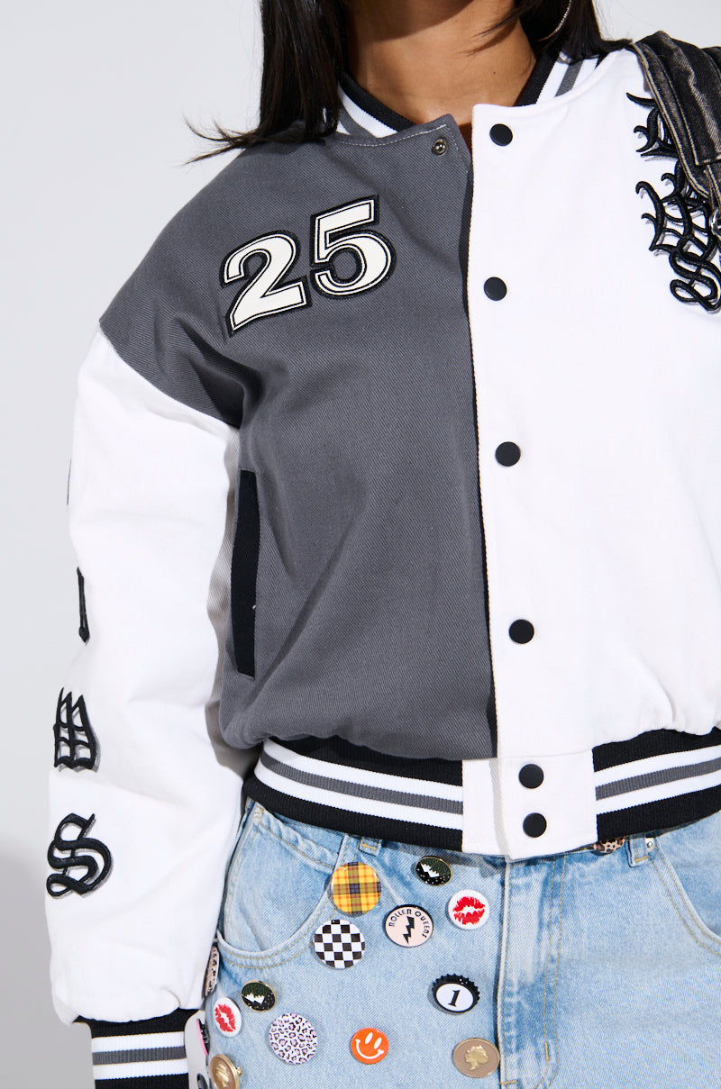 A person wears the NOT YOUR BABE EMBROIDERED BOMBER, a two-tone varsity jacket with "25" and sleeve patches. The grey-white design has striped cuffs. Paired with jeans decorated with colorful pins and patches, this outfit creates a vibrant look.