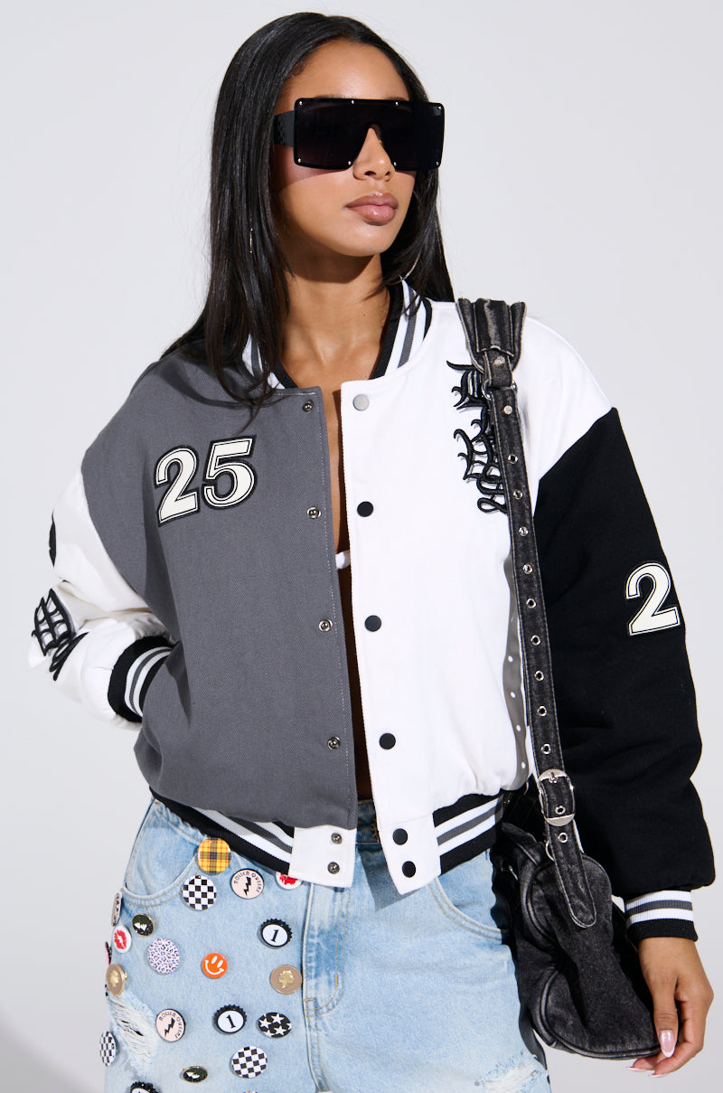 Wearing oversized sunglasses, a denim skirt with patches, and the NOT YOUR BABE EMBROIDERED BOMBER featuring "25" on the sleeves, the person carries a sleek black shoulder bag.