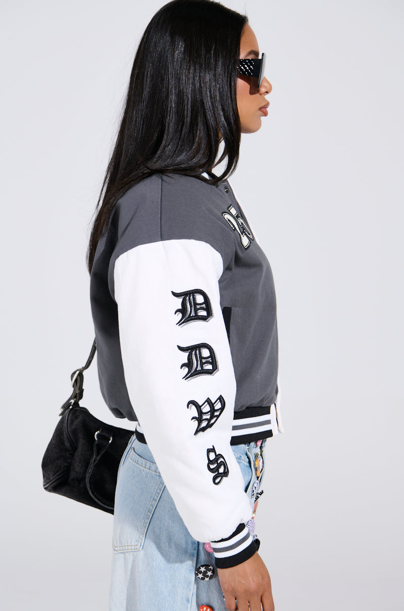 A person with long hair in sunglasses, wearing the NOT YOUR BABE EMBROIDERED BOMBER jacket adorned with black sleeve lettering, jeans featuring colorful patches, and carrying a black backpack, stands against a plain backdrop.