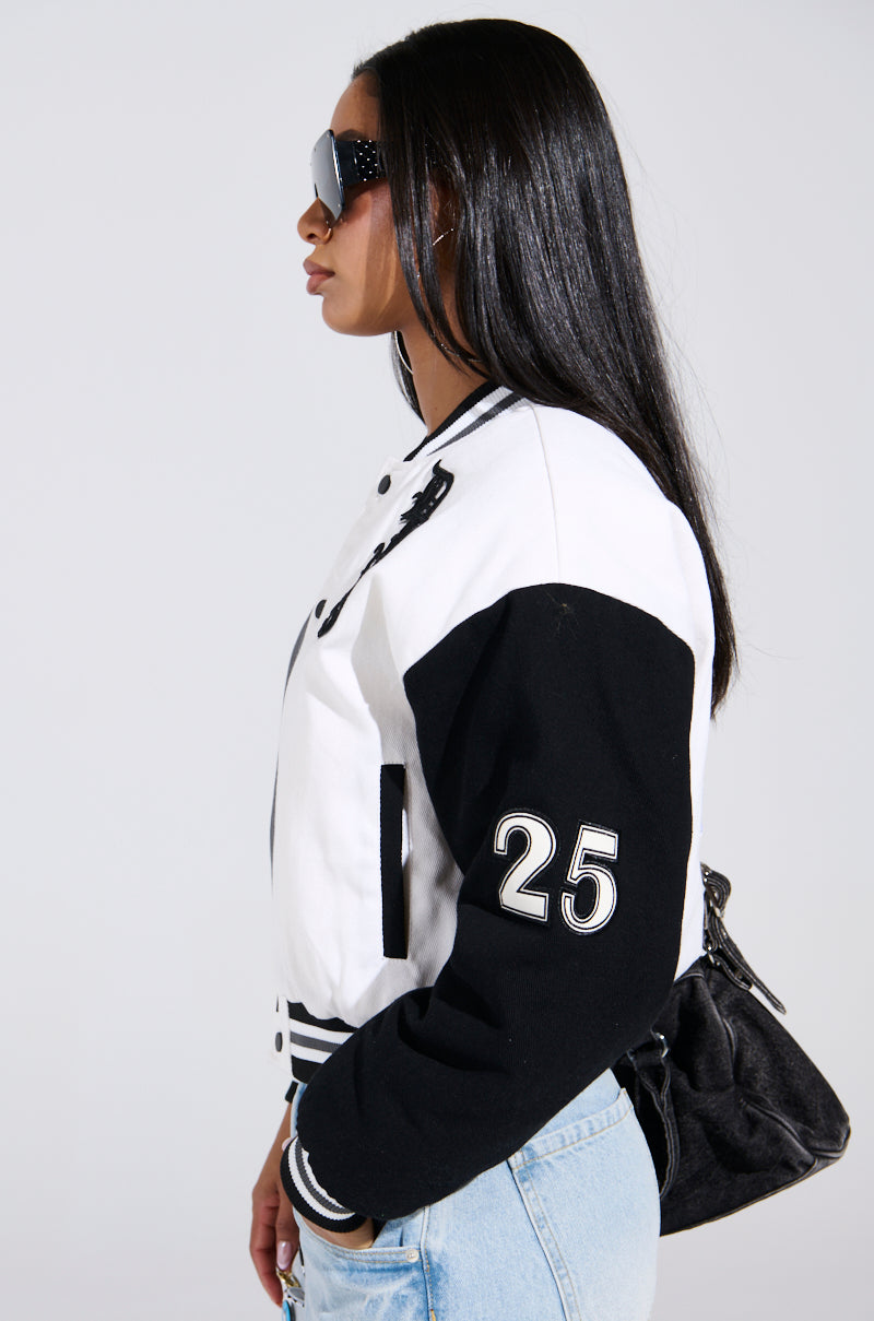 A woman with long black hair and sunglasses wears a NOT YOUR BABE EMBROIDERED BOMBER featuring the number 25 on the sleeve. She combines it with light blue jeans and a black bag, posing in profile against a plain backdrop.