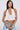 A woman with long dark hair wears the TIED TO YOU FRINGE TANK, a sleeveless white lace mesh cropped top, paired with light blue jeans, large hoop earrings, gold bracelets, and a white bag against a plain light background.