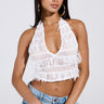 A woman with long dark hair wears the TIED TO YOU FRINGE TANK, a sleeveless white lace mesh cropped top, paired with light blue jeans, large hoop earrings, gold bracelets, and a white bag against a plain light background.