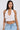 A woman with long dark hair wears the TIED TO YOU FRINGE TANK—a white, textured mesh cropped top—with light blue jeans. She accessorizes with hoop earrings and a bracelet, standing against a plain light background and looking at the camera.