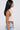 A woman with long straight black hair is shown in profile wearing the TIED TO YOU FRINGE TANK, a white crochet halter top, paired with light blue denim jeans against a plain light background.