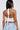 A woman with long black hair wears the TIED TO YOU FRINGE TANK, a white cropped halter top with a tie at the neck, paired with light blue jeans. Photographed from behind, the image showcases the tank’s backless design and her gold hoop earrings.