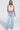 A woman exudes confidence in the TIED TO YOU FRINGE TANK, featuring a cropped white silhouette with fringe, paired with light blue wide-leg jeans. She accessorizes with a white clutch, open-toed sandals, and sleek hair for a chic look.