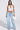 A woman stands confidently in the TIED TO YOU FRINGE TANK, a white mesh cropped top, paired with light blue high-waisted wide-leg jeans. She carries a white textured handbag, wears open-toe shoes, and her long hair flows freely.