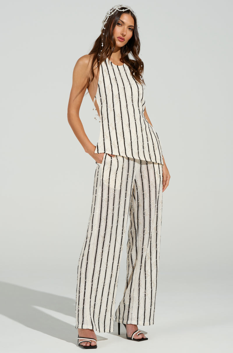 A woman models a sleeveless, backless white top with black vertical stripes, paired with the LOVELY DAYS KNIT PANT, featuring a matching wide-leg, high-rise fit. She completes the look with open-toed black and white sandals and a decorative headpiece in her long hair.