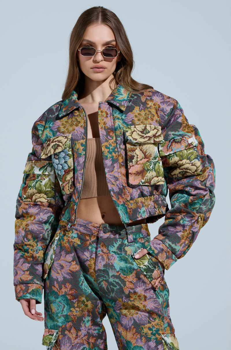 A woman in sunglasses dons the vibrant LOGAN SQUARE JACQUARD BOMBER, a tapestry-like floral jacket and pants set with oversized pockets, contrasting against a light blue background.