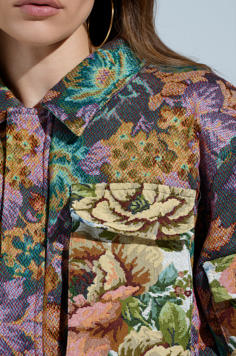 Close-up of a woman's upper body wearing the LOGAN SQUARE JACQUARD BOMBER, showcasing its floral pattern with vibrant green, yellow, and purple hues. The tapestry-like material features ruched detailing and a large pocket. She completes her look with hoop earrings.