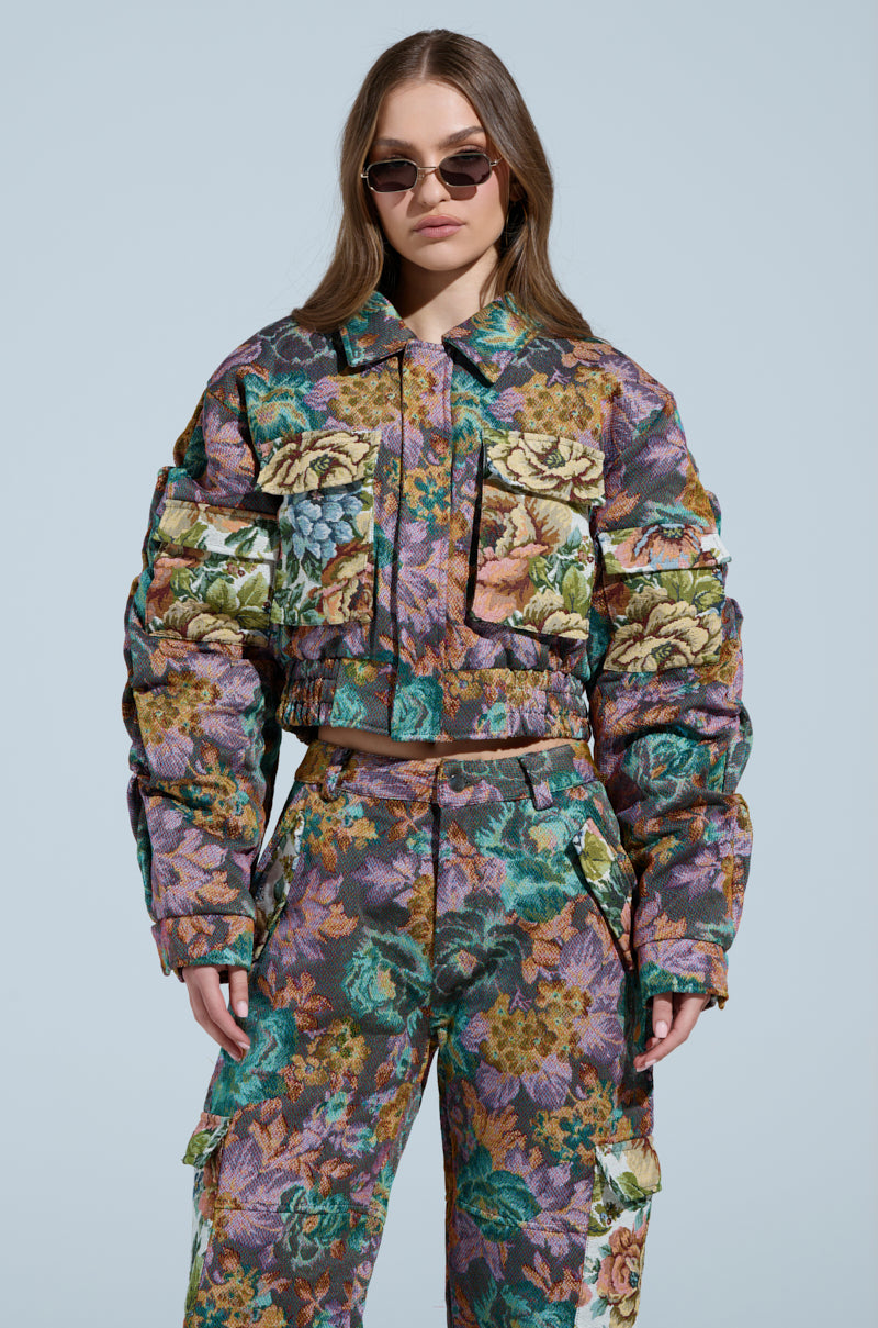 A person in a vibrant LOGAN SQUARE JACQUARD BOMBER, paired with matching pants and sunglasses, poses against a plain light blue background. The tapestry-like fabric enhances the floral pattern, exuding an effortlessly chic vibe.