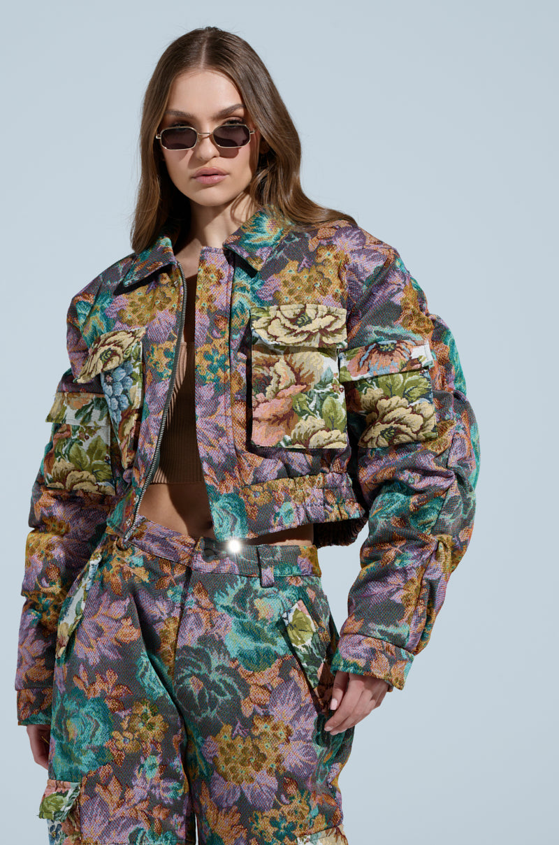 A woman confidently poses in sunglasses against a light blue background, donning a colorful tapestry-like jacket and matching ruched pants. Her long hair flows freely, embodying the style and composure reminiscent of the Logan Square Jacquard Bomber fashion vibe.